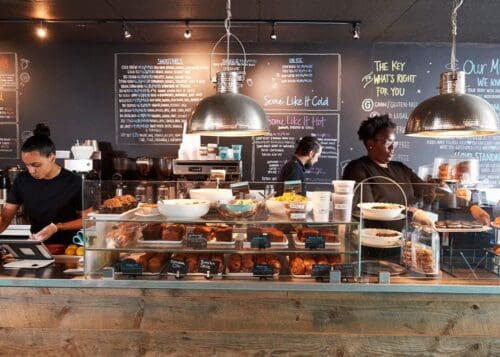 A cafe shopfront with workers and cabinet of food.