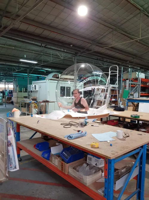 Person working in a workshop sitting inside a transparent bubble chair on a table, surrounded by tools, papers, and various equipment.